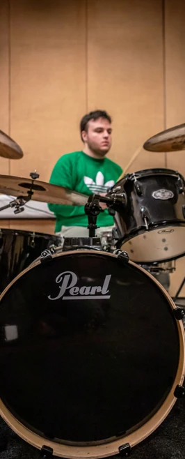A drummer wearing a green hoodie playing a Pearl drum kit in a soundproofed practice space. A drummer wearing a green hoodie playing a Pearl drum kit in a soundproofed practice space.